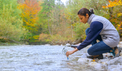 A person crouches by a river, measuring water quality with a device while surrounded by trees with autumn-colored leaves
