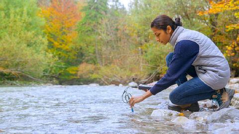 A person crouches by a river, measuring water quality with a device while surrounded by trees with autumn-colored leaves