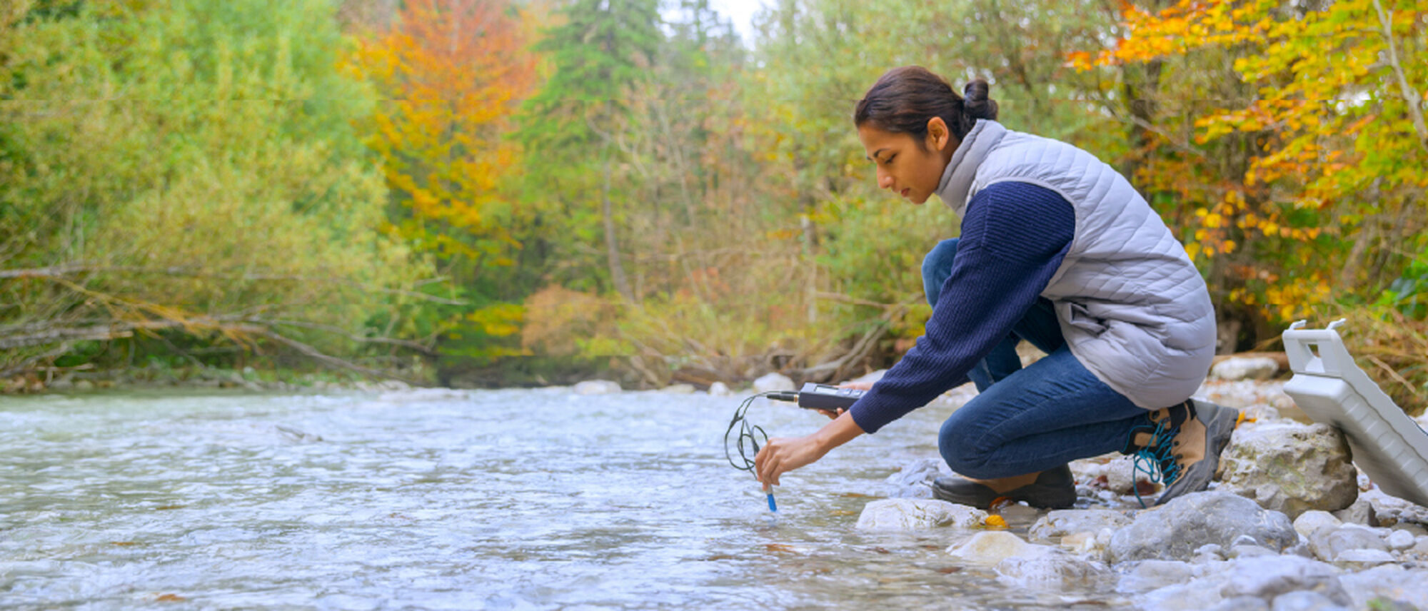 A person crouches by a river, measuring water quality with a device while surrounded by trees with autumn-colored leaves