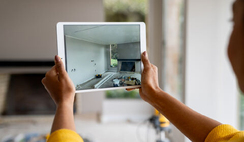 A person holds a tablet displaying a room under renovation, showing tools and materials on the floor