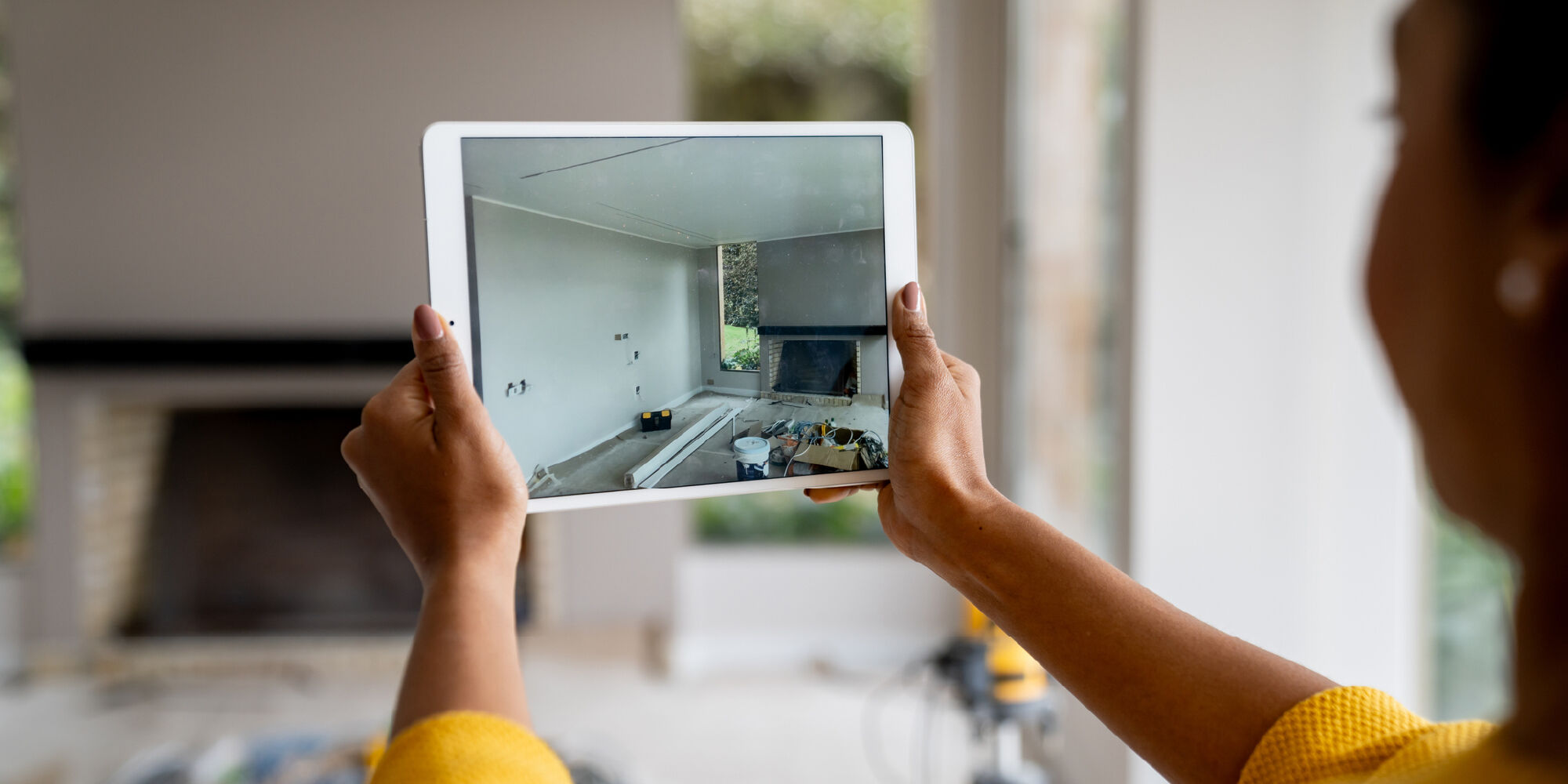 A person holds a tablet displaying a room under renovation, showing tools and materials on the floor
