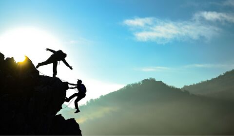 Two climbers assisting each other on a rocky cliff against a backdrop of a sunrise and misty mountains