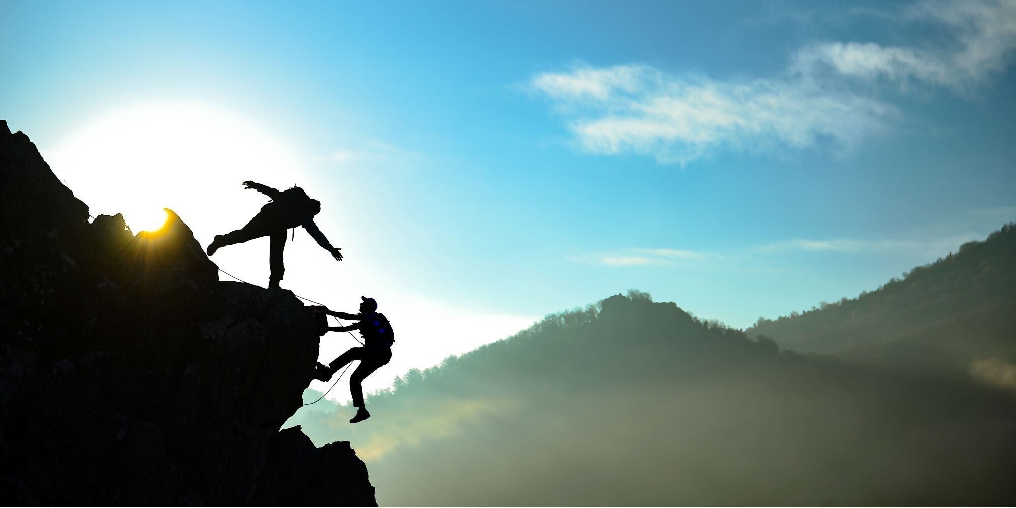 Two climbers assisting each other on a rocky cliff against a backdrop of a sunrise and misty mountains