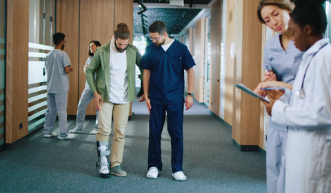A person with a leg brace stands with a healthcare worker in a hospital corridor, while others attend to patients nearby