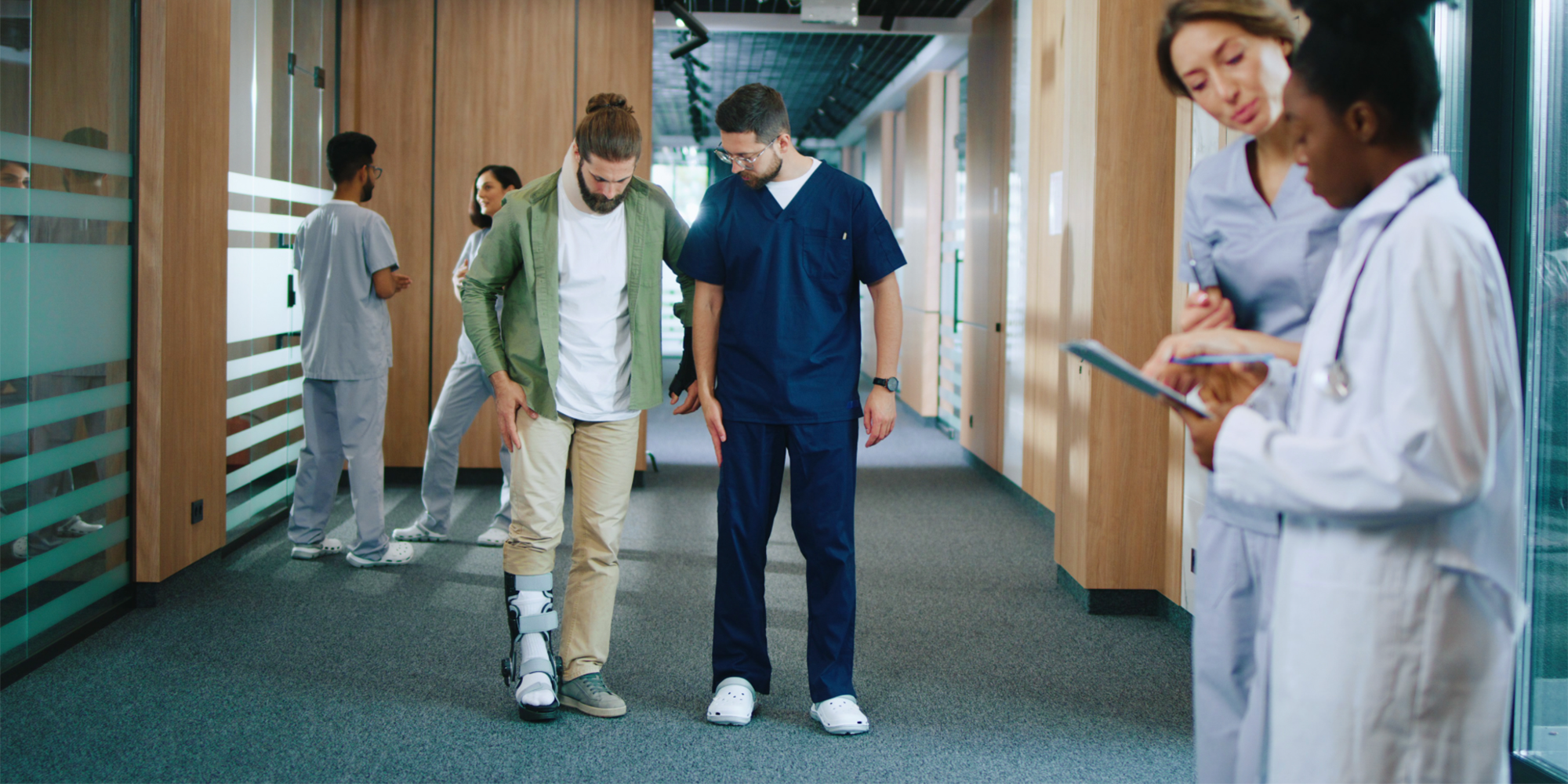 A person with a leg brace stands with a healthcare worker in a hospital corridor, while others attend to patients nearby
