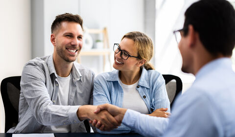 Two people shake hands at a table, smiling, while a third person observes. The setting is an office with natural light