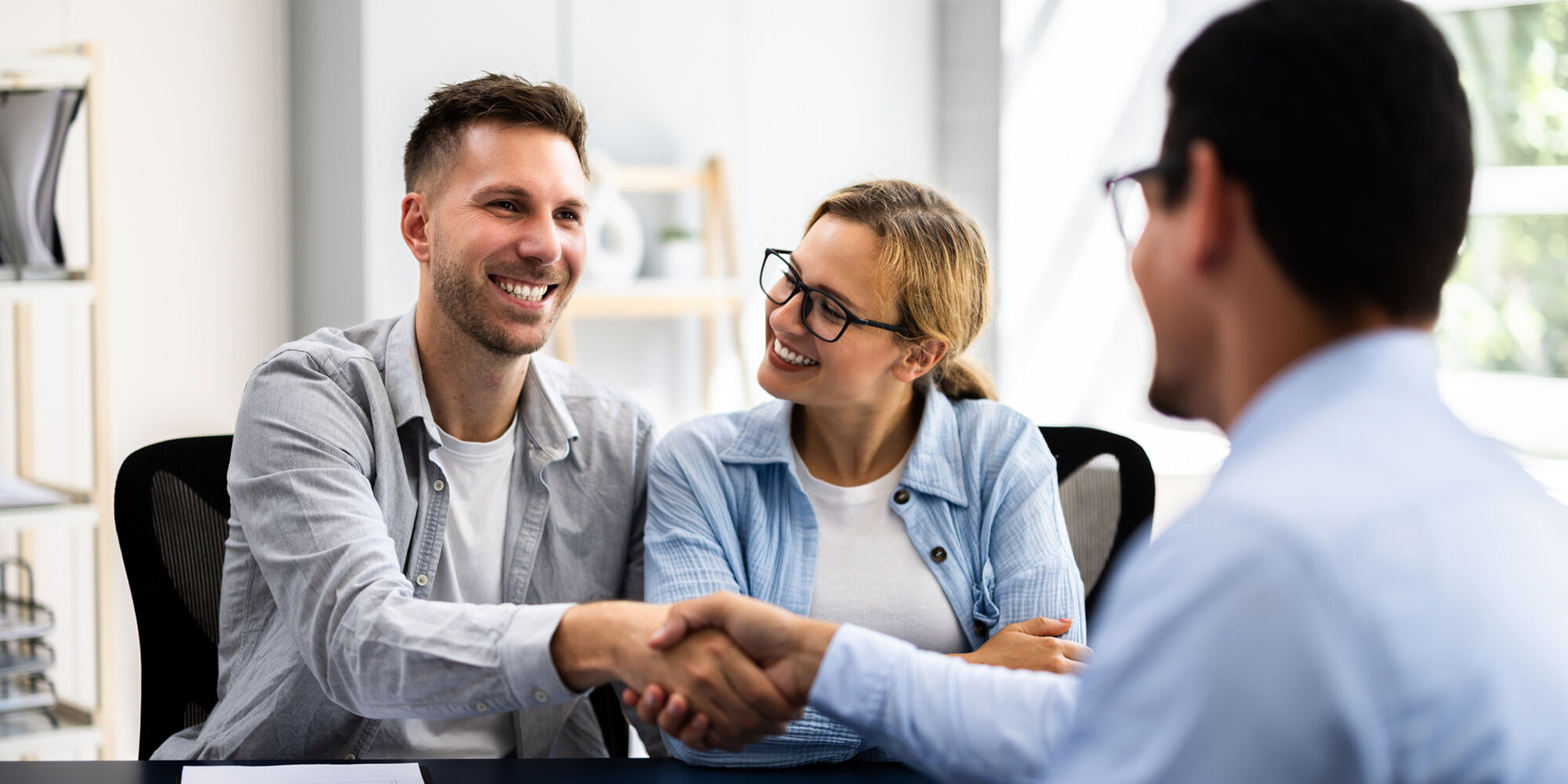 Two people shake hands at a table, smiling, while a third person observes. The setting is an office with natural light