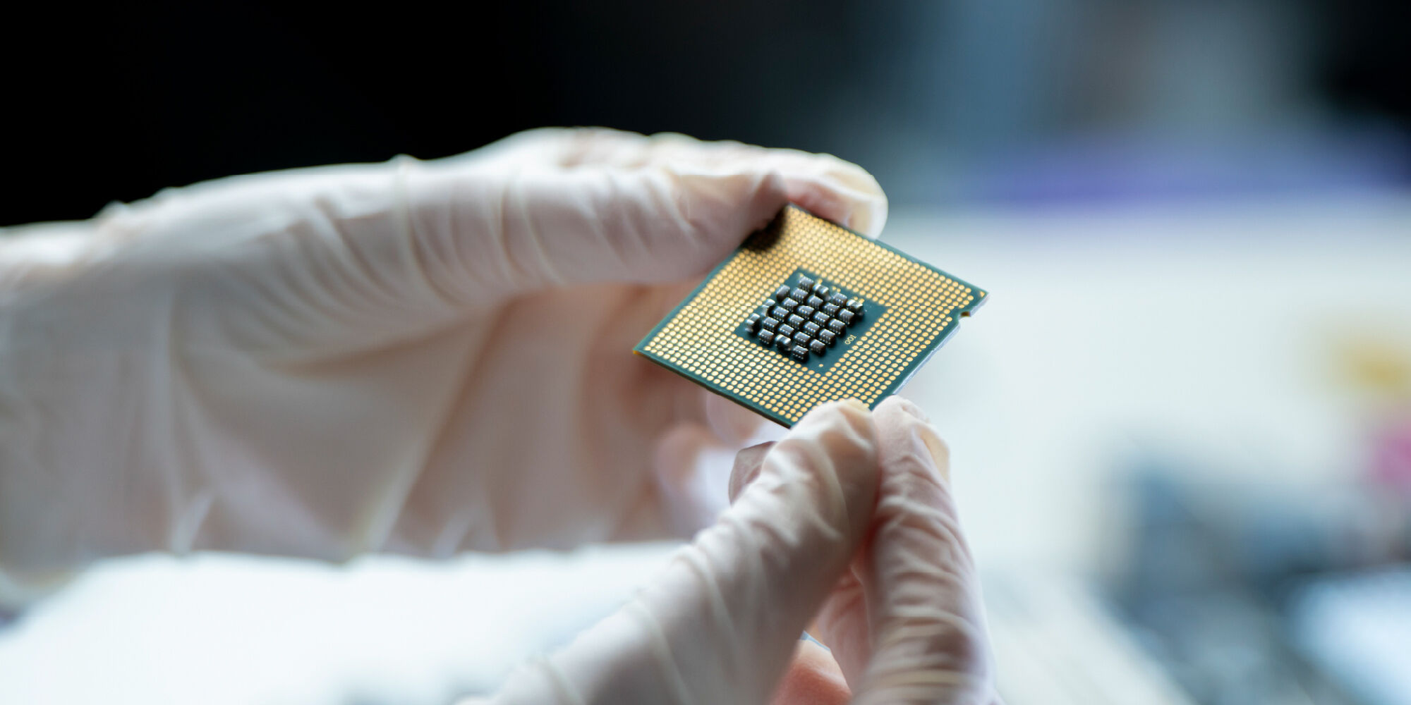 A person's hands in gloves holding a computer processor with a grid of pins on a reflective surface