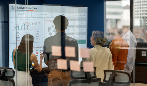 A group of people viewing a data analytics presentation on a large screen while discussing insights, with reflections on glass