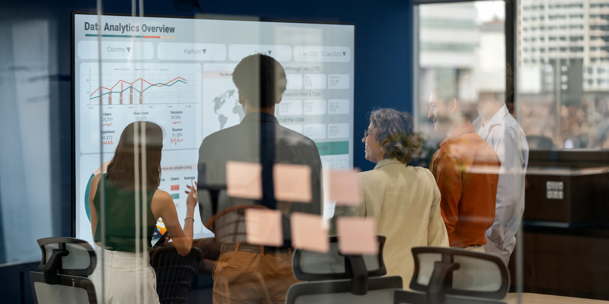 A group of people viewing a data analytics presentation on a large screen while discussing insights, with reflections on glass