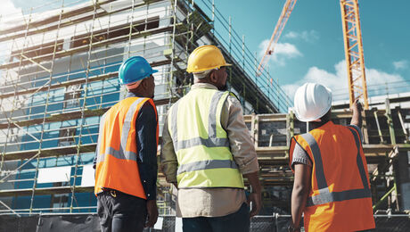 Construction workers in safety vests and hard hats reviewing a building site with scaffolding and cranes in the background.
