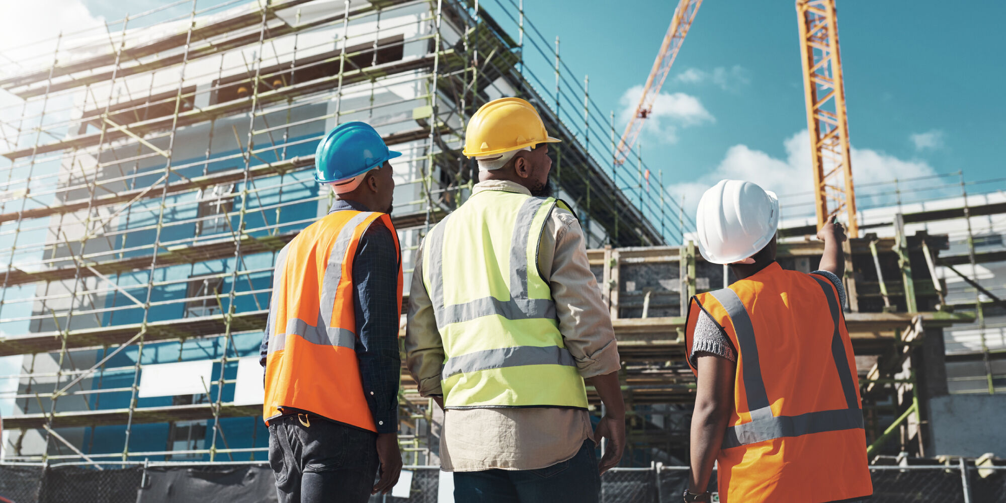 Construction workers in safety vests and hard hats reviewing a building site with scaffolding and cranes in the background.