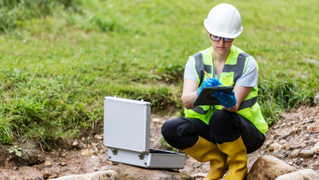 Environmental field inspector wearing safety gear uses a tablet beside monitoring equipment near a small stream.