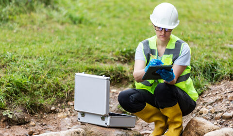 Environmental field inspector wearing safety gear uses a tablet beside monitoring equipment near a small stream.