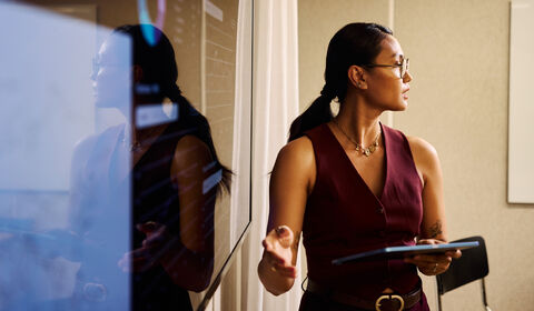 Person presenting beside a glass board, holding a tablet and gesturing, with reflections visible on the glass surface.