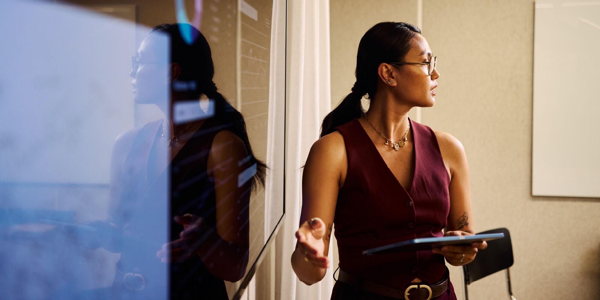 Person presenting beside a glass board, holding a tablet and gesturing, with reflections visible on the glass surface.
