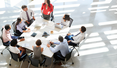A diverse group of eight people engaged in a meeting around a large table with laptops and documents in a bright, airy room