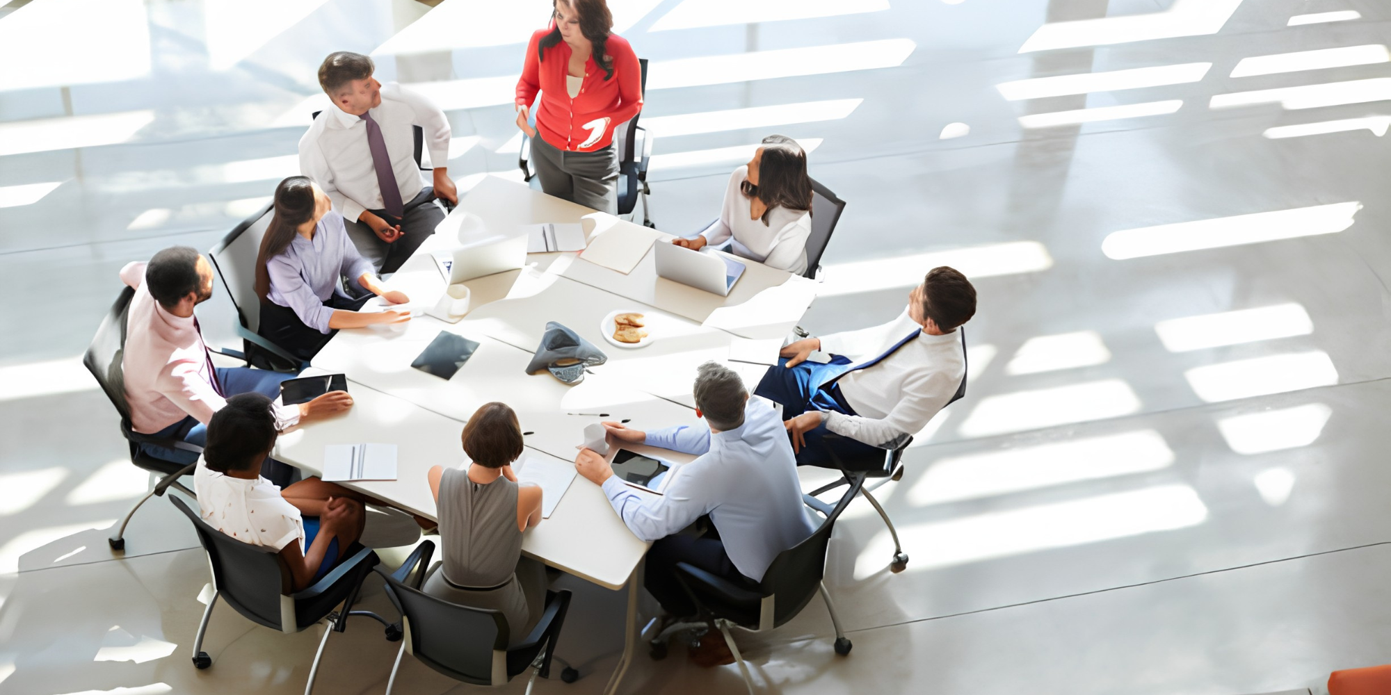 A diverse group of eight people engaged in a meeting around a large table with laptops and documents in a bright, airy room