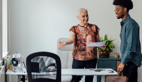 Person holding a document while extending a hand toward another person standing beside a desk in a bright office with a chair, small fan, and potted plant in view.