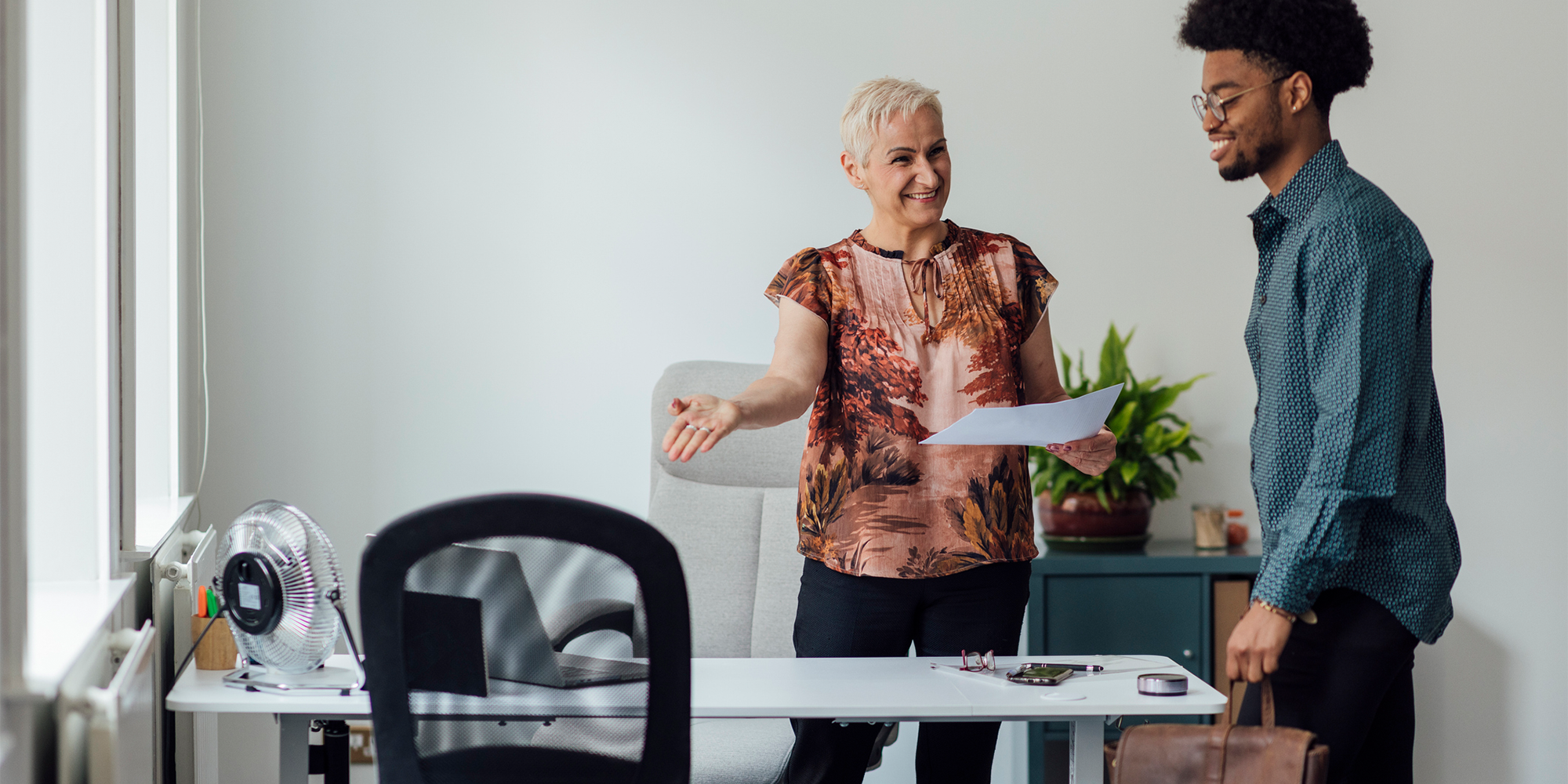 Person holding a document while extending a hand toward another person standing beside a desk in a bright office with a chair, small fan, and potted plant in view.