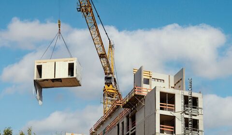 A construction crane lifts a building module towards a partially completed concrete structure under a blue sky with clouds
