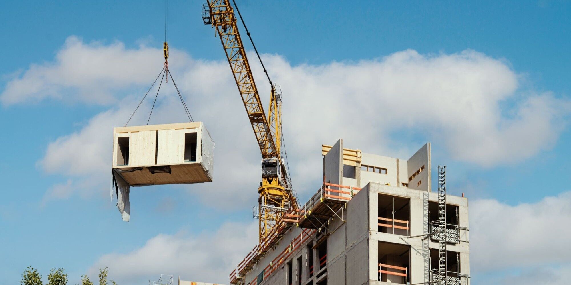 A construction crane lifts a building module towards a partially completed concrete structure under a blue sky with clouds