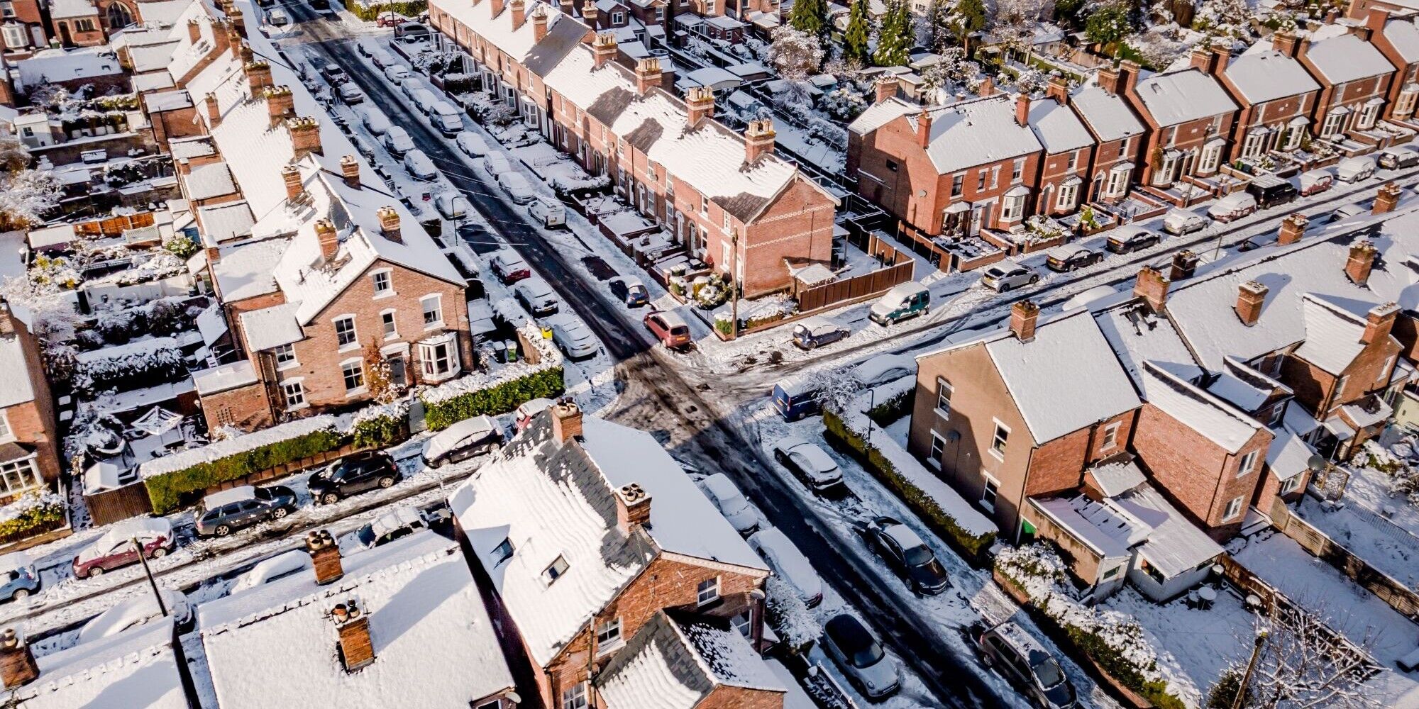A snowy aerial view of a residential neighborhood with rows of brick houses and cars lining the streets.