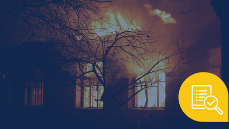 A building engulfed in flames at night, with bright orange fire contrasting against the dark silhouette of nearby trees