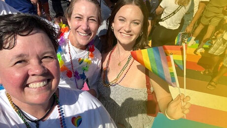 A group of three individuals smiling at a pride event, wearing colorful accessories, with one holding a rainbow flag