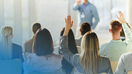 A group of people seated in a conference room, with some raising their hands, while another person is speaking in front