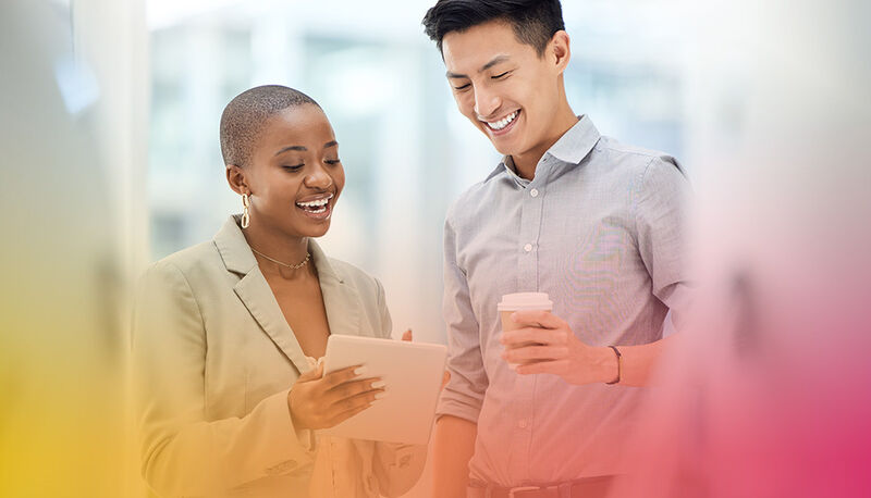 Two individuals share a light moment while looking at a tablet, one holding a coffee cup, in a bright, softly blurred setting