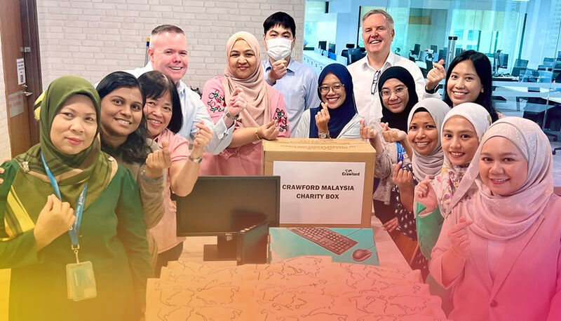 A diverse group of people poses around a charity box labeled "Crawford Malaysia Charity Box," smiling and giving thumbs up