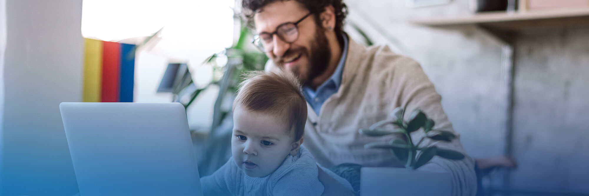 A person with curly hair and glasses smiles while using a laptop, with a baby crawling nearby and a plant in the background