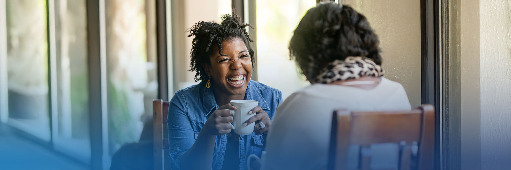 Two individuals are seated indoors, engaged in a joyful conversation while holding coffee mugs