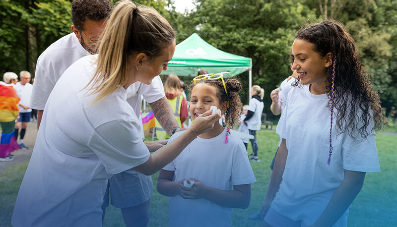 A person applies face paint to a smiling child, while two others watch, all in casual white shirts at an outdoor event
