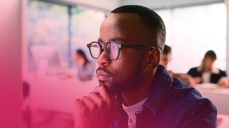 A person with glasses and a beard rests their chin on their hand, deep in thought, in a classroom setting with blurred figures in the background