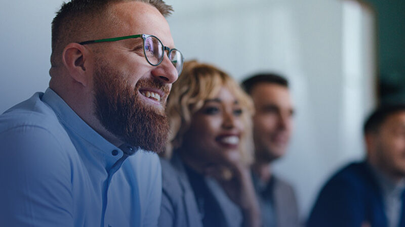 A group of people in a meeting, smiling and engaged, with a focus on one individual with glasses and a beard