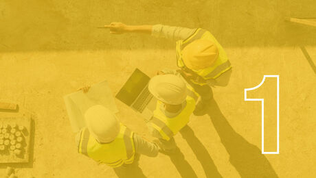 Three construction workers in yellow vests and helmets collaborate on plans with a laptop, one gesturing while holding a pen