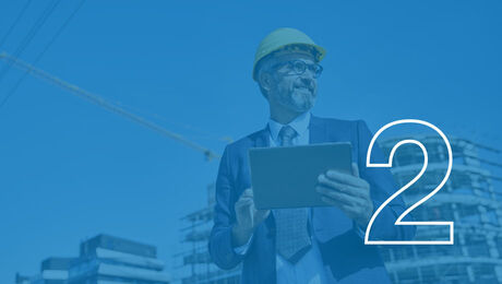 A person in a suit and hard hat holds a tablet, standing in front of a construction site with cranes in the background
