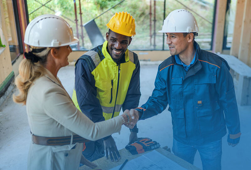 Three construction professionals in hard hats shake hands over a project plan inside a partially built structure