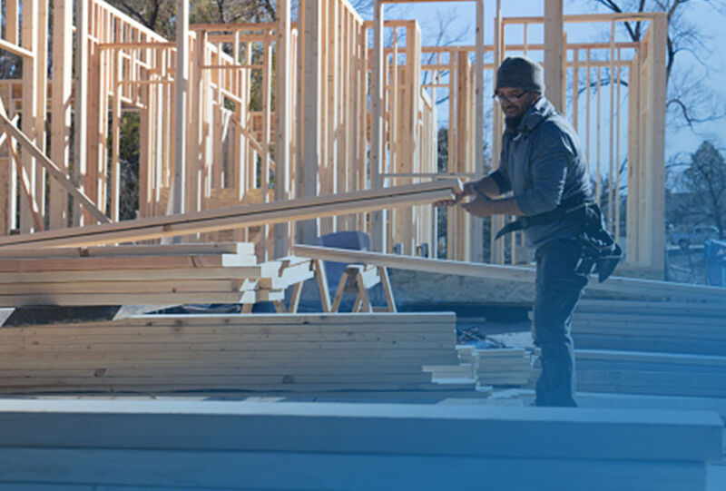 A person is lifting wooden planks at a construction site with a partially framed structure in the background