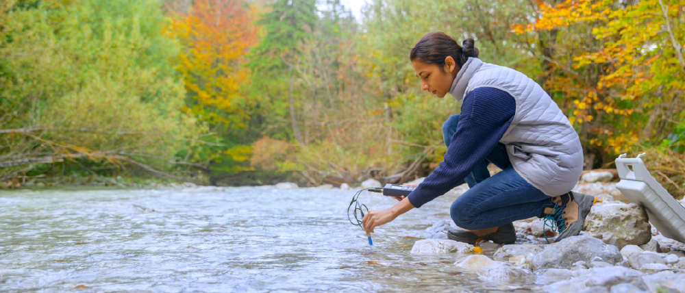 A person crouches by a river, measuring water quality with a device while surrounded by trees with autumn-colored leaves