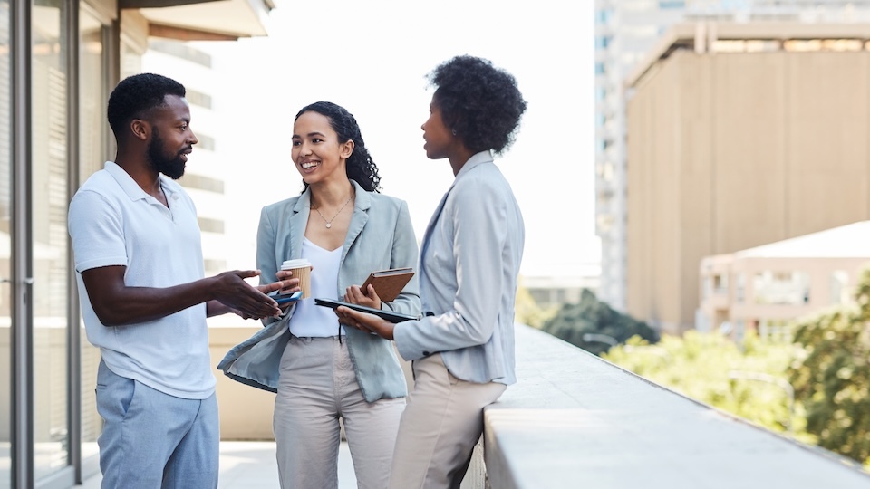 Three individuals converse on a balcony, one holding a coffee cup, another with a tablet, and the third with a folder, surrounded by urban scenery